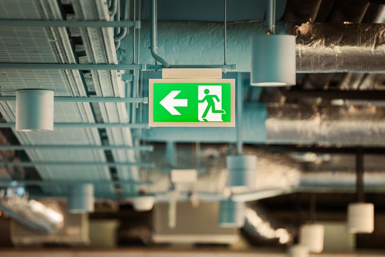 Green emergency exit sign hanging from an industrial ceiling, directing towards safety.